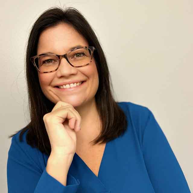 A smilling women, wearing transparent glasses and her hand on her chin.